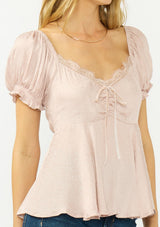 [Color: Dusty Rose] A detail image of a blonde model wearing a pink satin jacquard puff sleeve top with lace trim and a tie front detail. 