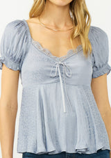 [Color: Dusty Blue] A detail image of a blonde model wearing a blue satin jacquard puff sleeve top with lace trim and a tie front detail. 