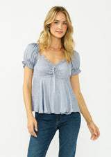 [Color: Dusty Blue] A front facing image of a blonde model wearing a blue satin jacquard puff sleeve top with lace trim and a tie front detail. 