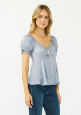 [Color: Dusty Blue] A side facing image of a blonde model wearing a blue satin jacquard puff sleeve top with lace trim and a tie front detail. 