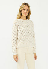 [Color: Natural] A side facing image of a blonde model wearing a cozy off white pointelle knit sweater with a crew neckline. 
