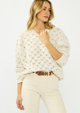 [Color: Natural] A front facing image of a blonde model wearing a cozy off white pointelle knit sweater with a crew neckline. Tucked into a beige denim with a brown belt. 