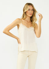[Color: Champagne] A front facing image of a blonde model wearing an ivory satin camisole with lace trim, adjustable spaghetti straps, and side slits. 