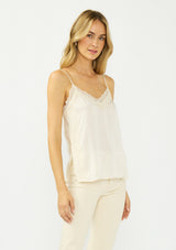 [Color: Champagne] A side facing image of a blonde model wearing an ivory satin camisole with lace trim, adjustable spaghetti straps, and side slits. 