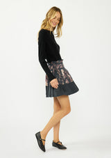[Color: Grey/Dusty Rose] A side facing image of a blonde model wearing a grey floral pleated skirt with a satin hem, clipped lurex, paperbag waist, and side tie details. 