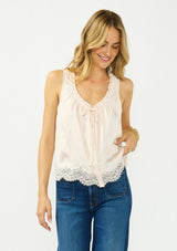 [Color: Petal] A front facing image of a blonde model wearing an light pink poly satin tank top with a ruffled v neckline, tie front, button front, and a lace trim hem. 