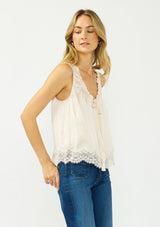 [Color: Petal] A side facing image of a blonde model wearing an light pink poly satin tank top with a ruffled v neckline, tie front, button front, and a lace trim hem. 