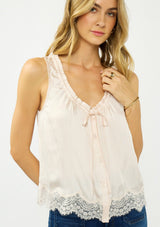 [Color: Petal] A front facing image of a blonde model wearing an light pink poly satin tank top with a ruffled v neckline, tie front, button front, and a lace trim hem. 