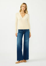 [Color: Cream] A front facing image of a blonde model wearing a cream surplice sweater with a ribbed hem and side button closure. 