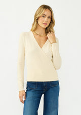 [Color: Cream] A front facing image of a blonde model wearing a cream surplice sweater with a ribbed hem and side button closure. 