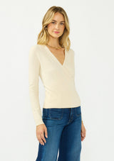 [Color: Cream] A side facing image of a blonde model wearing a cream surplice sweater with a ribbed hem and side button closure. 