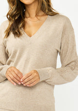 [Color: Heather Taupe] A detail image of a brunette model wearing a beige v neck sweater with a cable knit detail and styled with a matching bottom. 