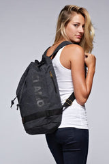 [Color: Black] A blonde model wearing a cotton blend black sack backpack with a drawstring closure. 