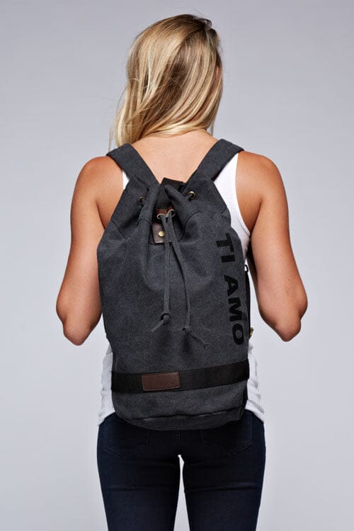 [Color: Black] A blonde model wearing a cotton blend black sack backpack with a drawstring closure. 
