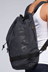 [Color: Black] A blonde model wearing a cotton blend black sack backpack with a drawstring closure. 