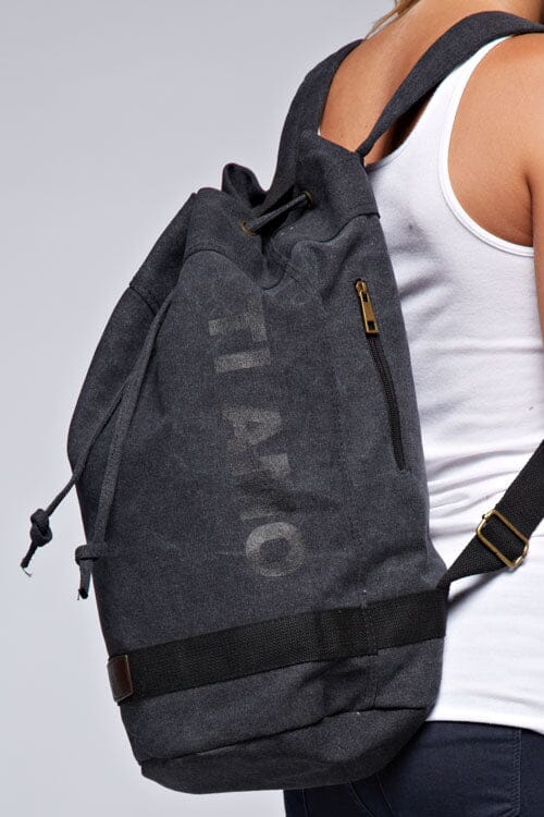 [Color: Black] A blonde model wearing a cotton blend black sack backpack with a drawstring closure. 