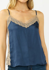 [Color: Navy/Taupe] A detail image of a blonde model wearing a navy satin camisole with lace trim, adjustable spaghetti straps, and side slits. 