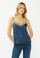[Color: Navy/Taupe] A front facing image of a blonde model wearing a navy satin camisole with lace trim, adjustable spaghetti straps, and side slits. 