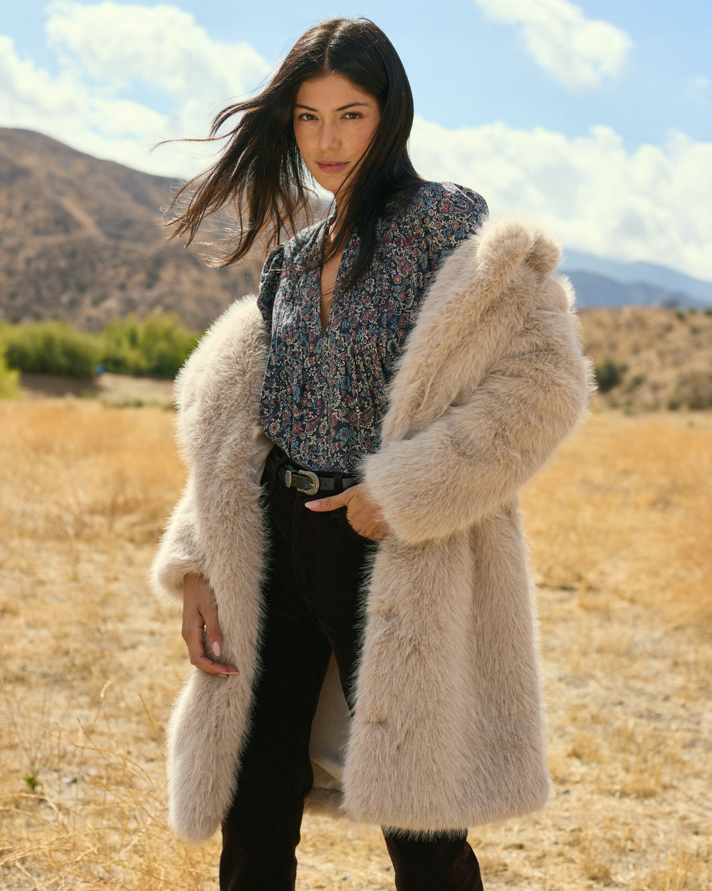 Woman wearing a fluffy beige coat in an outdoor setting with mountains in the background