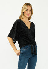 [Color: Black] A side facing image of a blonde model wearing a black floral burnout tie front top with flowy half length sleeves and a v neckline. 