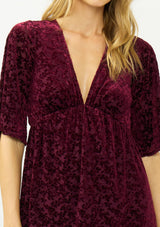 [Color: Merlot] A detail image of a blonde model wearing a burgundy velvet burnout mini dress with half sleeves, a v neckline, and an elastic empire waist. 