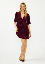 [Color: Merlot] A front facing image of a blonde model wearing a burgundy velvet burnout mini dress with half sleeves, a v neckline, and an elastic empire waist. 