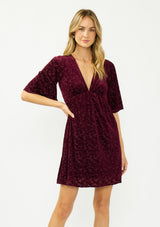 [Color: Merlot] A front facing image of a blonde model wearing a burgundy velvet burnout mini dress with half sleeves, a v neckline, and an elastic empire waist. 
