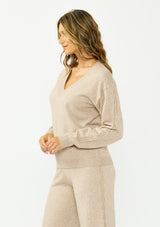 [Color: Heather Taupe] A side facing image of a brunette model wearing a beige v neck sweater with a cable knit detail and styled with a matching bottom. 