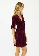 [Color: Merlot] A side facing image of a blonde model wearing a burgundy velvet burnout mini dress with half sleeves, a v neckline, and an elastic empire waist. 
