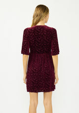 [Color: Merlot] A back facing image of a blonde model wearing a burgundy velvet burnout mini dress with half sleeves, a v neckline, and an elastic empire waist. 