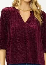 [Color: Merlot] A detail image of a blonde model wearing a burgundy floral burnout blouse with a v neckline three quarter length sleeve. 