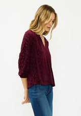 [Color: Merlot] A side facing image of a blonde model wearing a burgundy floral burnout blouse with a v neckline three quarter length sleeve. 