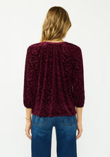 [Color: Merlot] A back facing image of a blonde model wearing a burgundy floral burnout blouse with a v neckline three quarter length sleeve. 