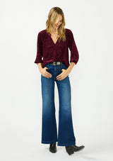 [Color: Merlot] A front facing image of a blonde model wearing a burgundy floral burnout blouse with a v neckline three quarter length sleeve. Styled tucked into a dark wash denim with a black belt and boots. 