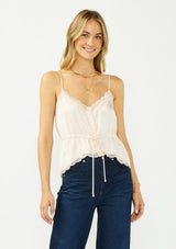 [Color: Antique Blush] A front facing image of a blonde model wearing a light pink poly satin lace trim top with  a button front, adjustable spaghetti straps, and a drawstring waist with tie detail. Paired with a classic dark wash denim. 