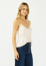 [Color: Antique Blush] A side facing image of a blonde model wearing a light pink poly satin lace trim top with  a button front, adjustable spaghetti straps, and a drawstring waist with tie detail. Paired with a classic dark wash denim. 
