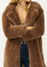 [Color: Mocha] A detail image of a blonde model wearing a brown faux fur coat with a shawl collared neckline, and side pockets. A large fur coat for the winter, holidays, and other special occasions. 