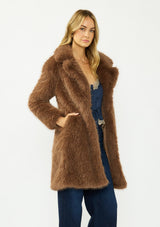 [Color: Mocha] A side facing image of a blonde model wearing a brown faux fur coat with a shawl collared neckline, and side pockets. A large fur coat for the winter, holidays, and other special occasions. 