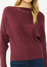 [Color: Oxblood] A detail image of a blonde model wearing a burgundy pullover sweater with a boat neckline and long sleeves that taper at the cuffs. 