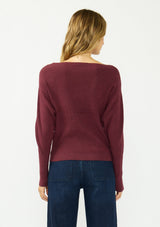[Color: Oxblood] A back facing image of a blonde model wearing a burgundy pullover sweater with a boat neckline and long sleeves that taper at the cuffs. 