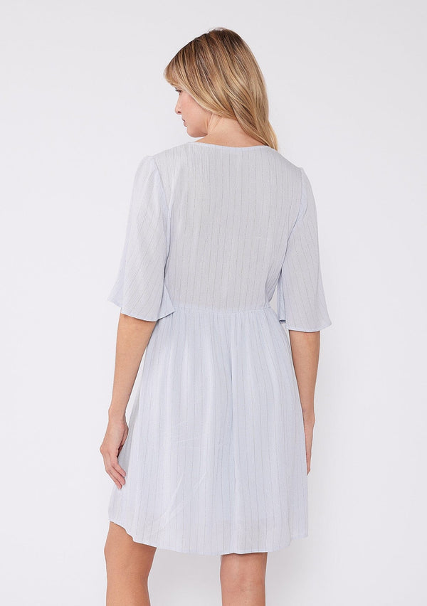 [Color: Periwinkle] A back facing image of a blonde model wearing a classic flowy mini dress in light blue with a gold metallic thread detail. With short half length sleeves, a v neckline, and an empire waist.