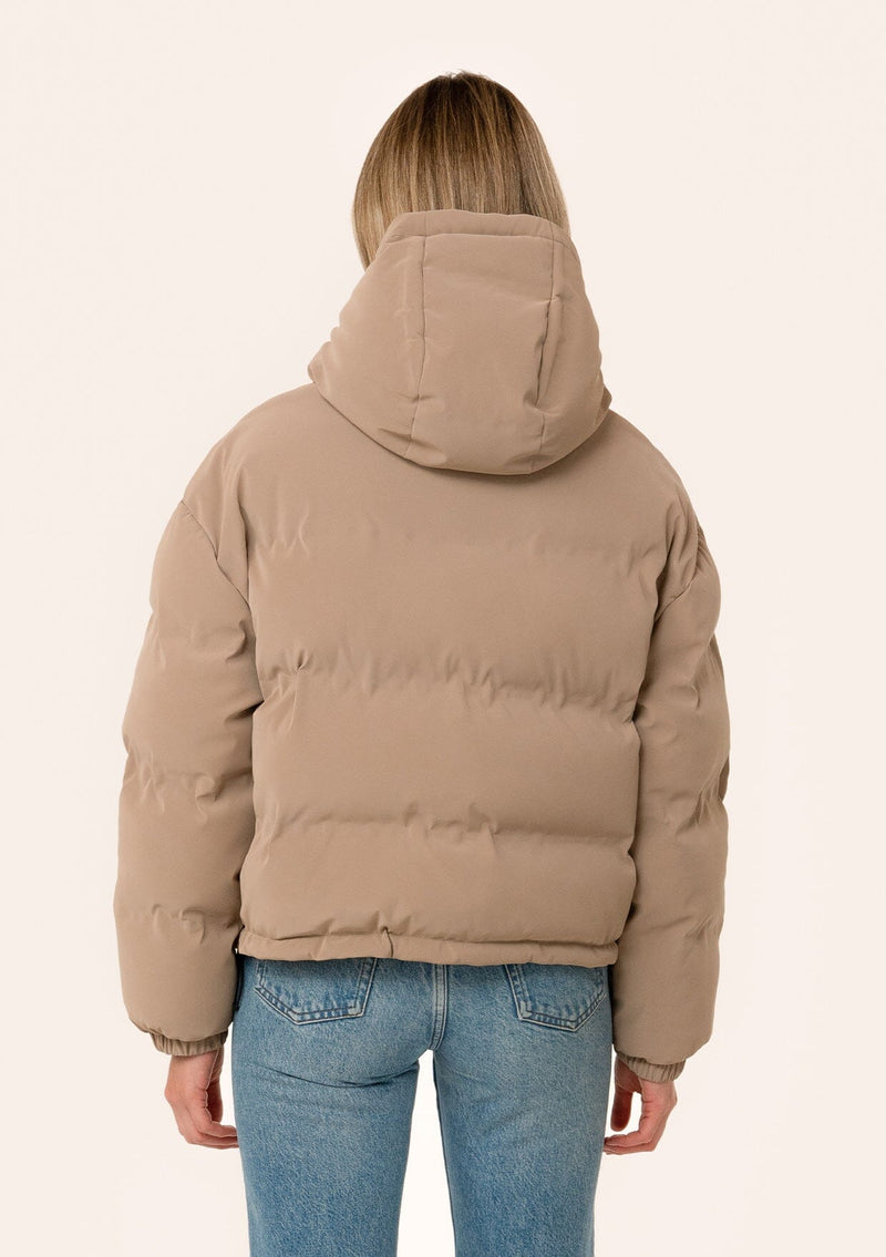 [Color: Taupe] A back facing image of a blonde model wearing an ultra puffy cropped jacket in a matte taupe brown finish. Featuring an adjustable hoodie with toggles, a zippered front, and side pockets. 