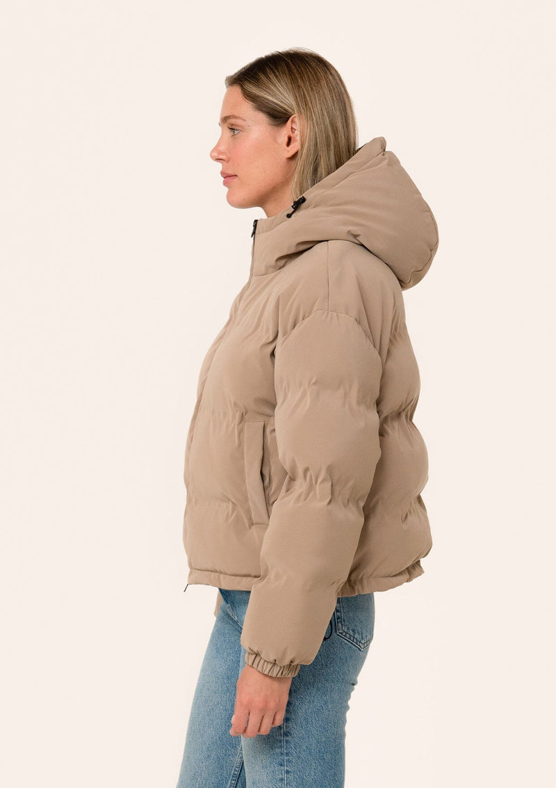 [Color: Taupe] A side facing image of a blonde model wearing an ultra puffy cropped jacket in a matte taupe brown finish. Featuring an adjustable hoodie with toggles, a zippered front, and side pockets. 