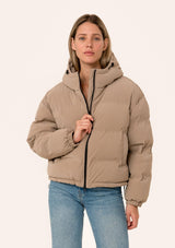 [Color: Taupe] A front facing image of a blonde model wearing an ultra puffy cropped jacket in a matte taupe brown finish. Featuring an adjustable hoodie with toggles, a zippered front, and side pockets. 