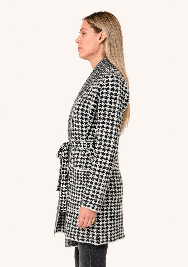 [Color: White/Black] A side facing image of a blonde model wearing a classic fall cardigan in a white and black houndstooth. With long sleeves, a shawl collar, side pockets, and a tie waist belt. 