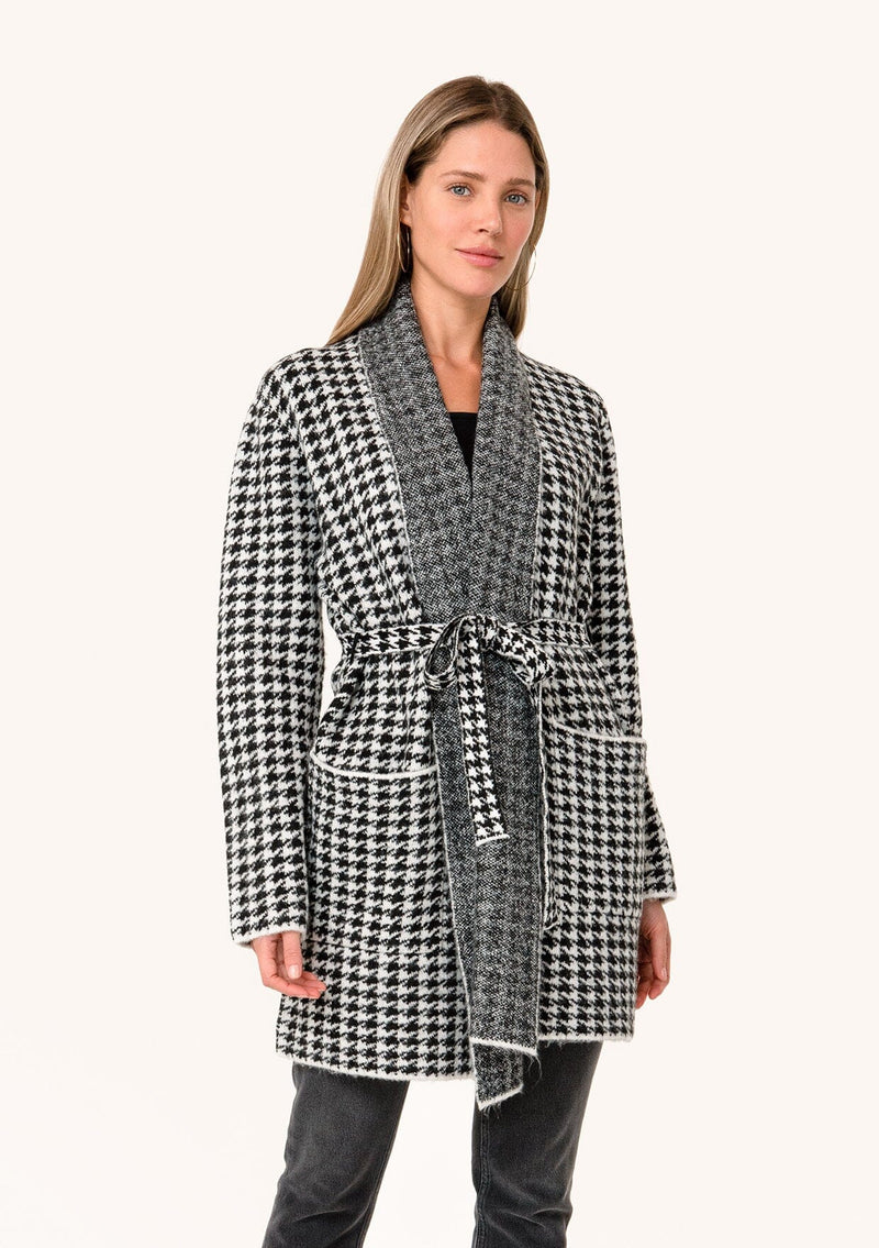 [Color: White/Black] A front facing image of a blonde model wearing a classic fall cardigan in a white and black houndstooth. With long sleeves, a shawl collar, side pockets, and a tie waist belt. 
