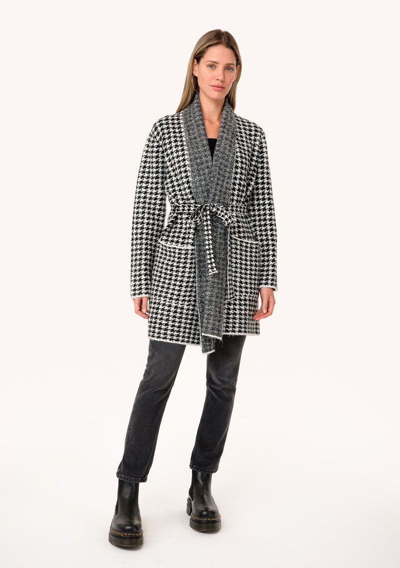 [Color: White/Black] A full body front facing image of a blonde model wearing a classic fall cardigan in a white and black houndstooth. With long sleeves, a shawl collar, side pockets, and a tie waist belt. 