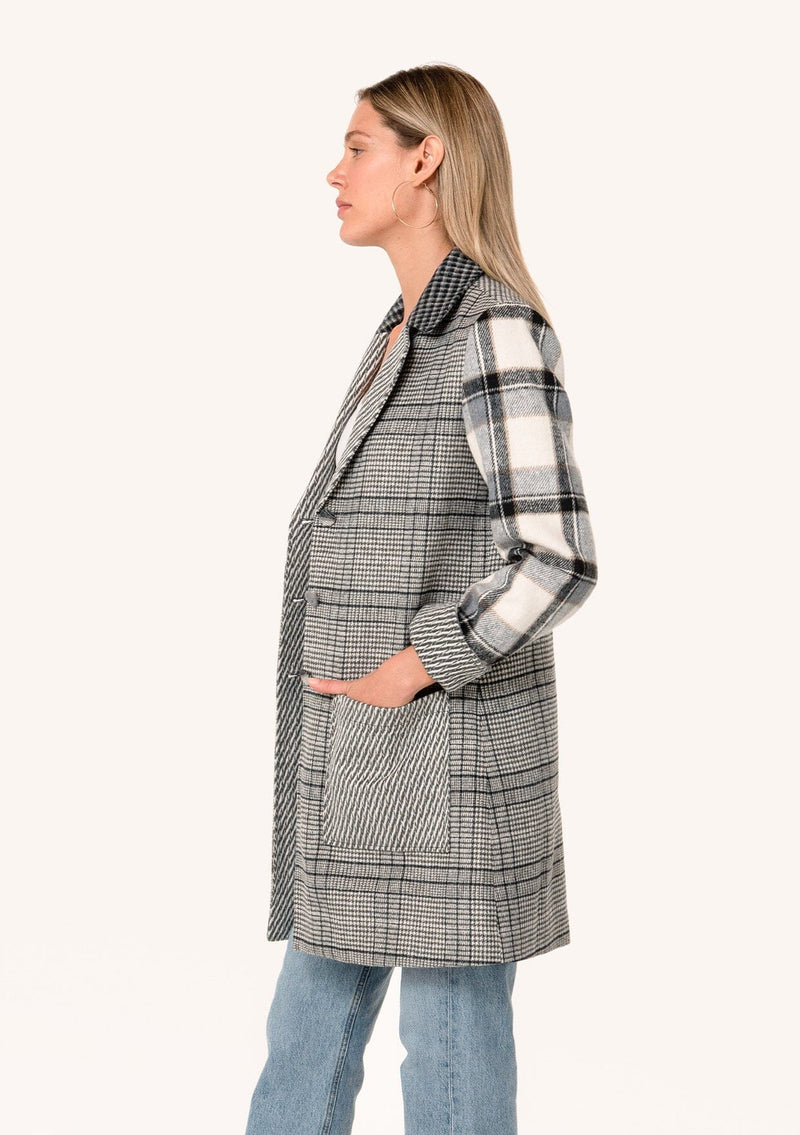 [Color: Taupe/Grey] A side facing image of a blonde model wearing a bohemian long blazer coat in a grey patchwork plaid. With long sleeves, a notched lapel, a button front, and patch side pockets. 