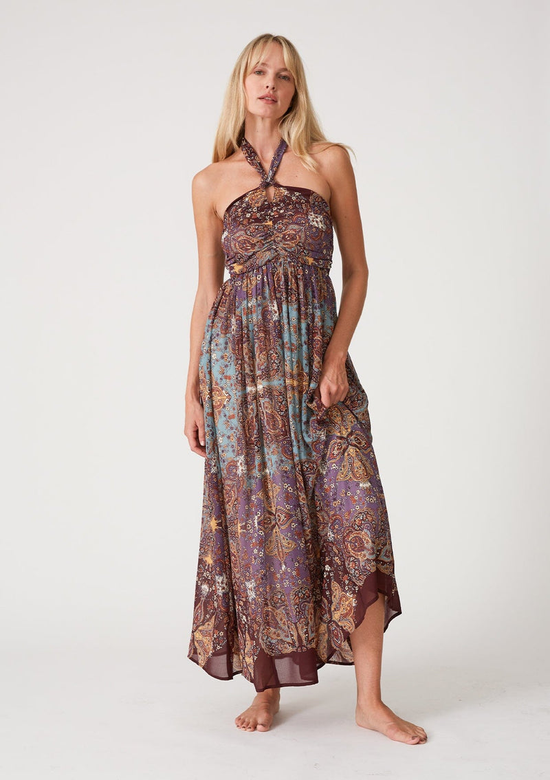 [Color: Raisin/Teal] A front facing image of a blonde model wearing a bohemian halter maxi dress in a multi colored purple and teal floral print. With a tie neckline, a slim fit bodice with a smocked elastic back, and a long flowy skirt.