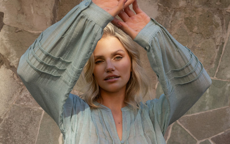 Woman wearing a light blue blouse with long sleeves against a stone wall.